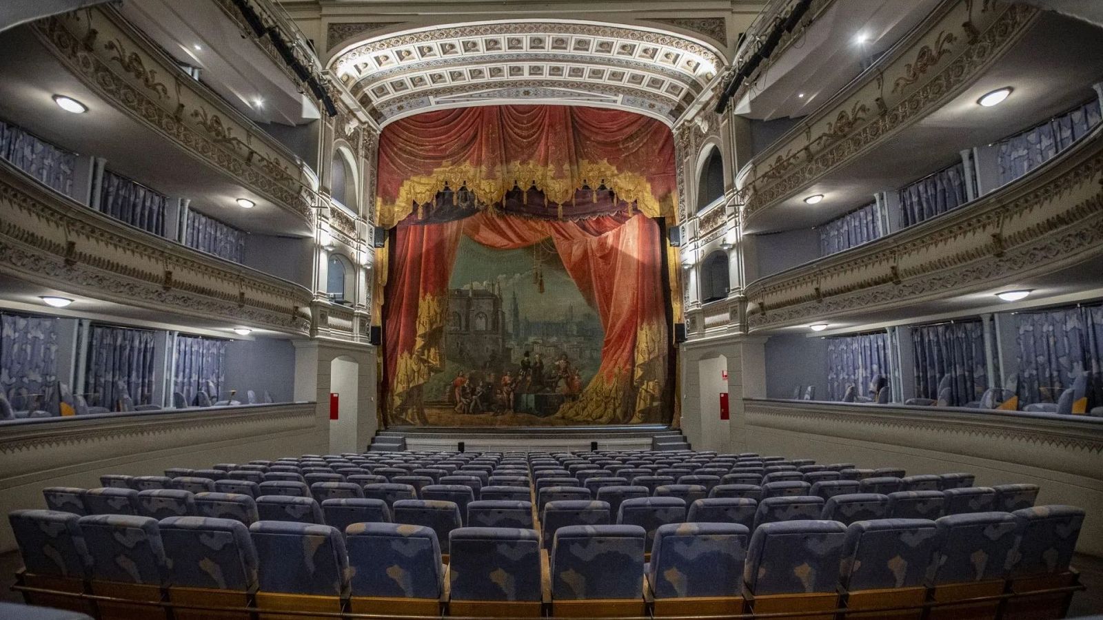 Vista interior de un teatro histórico con butacas azules, un elaborado telón rojo en el escenario y palcos laterales iluminados.