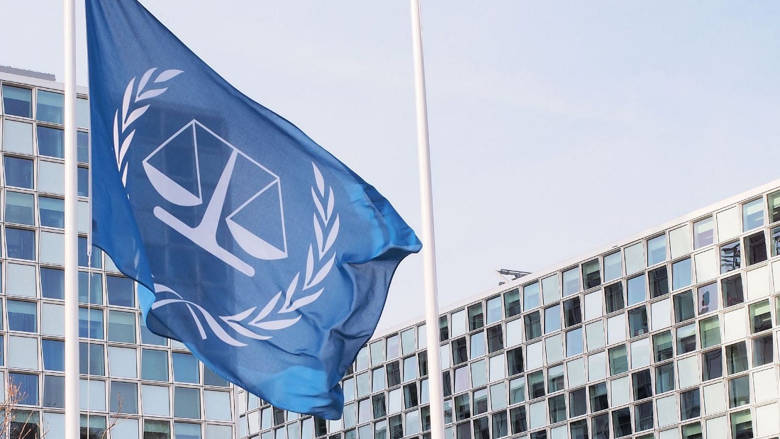 Bandera azul de la Corte Penal Internacional con emblema de balanza y ramas de olivo, ondeando frente a su sede moderna bajo un cielo despejado.