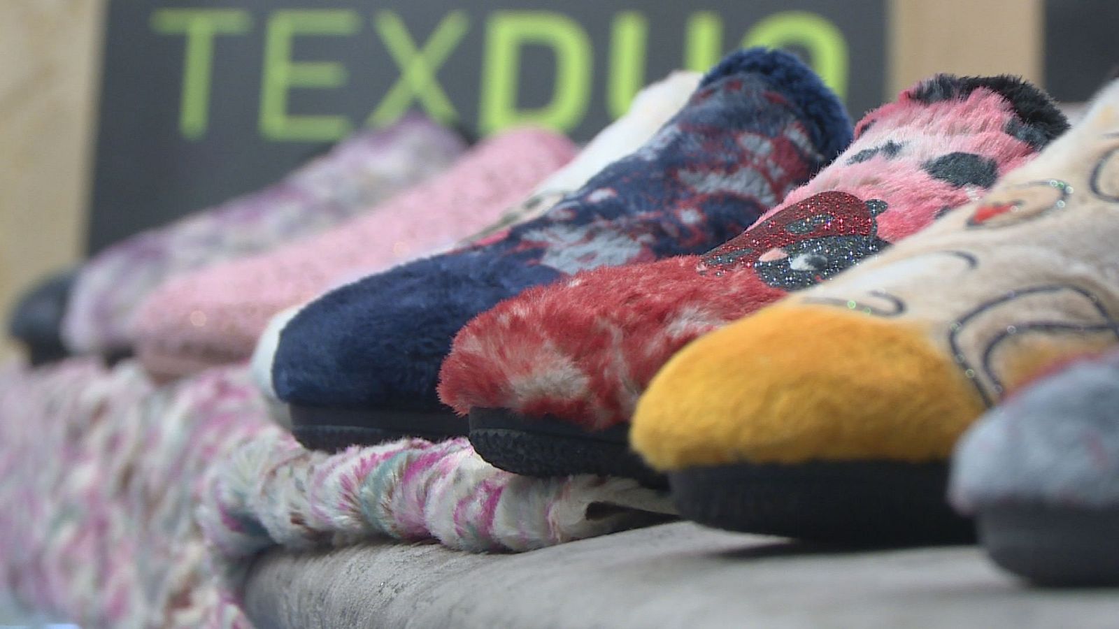 Hilera de zapatillas de invierno de suave tejido y diversos colores expuestas en diagonal sobre un soporte en una feria del sector.
