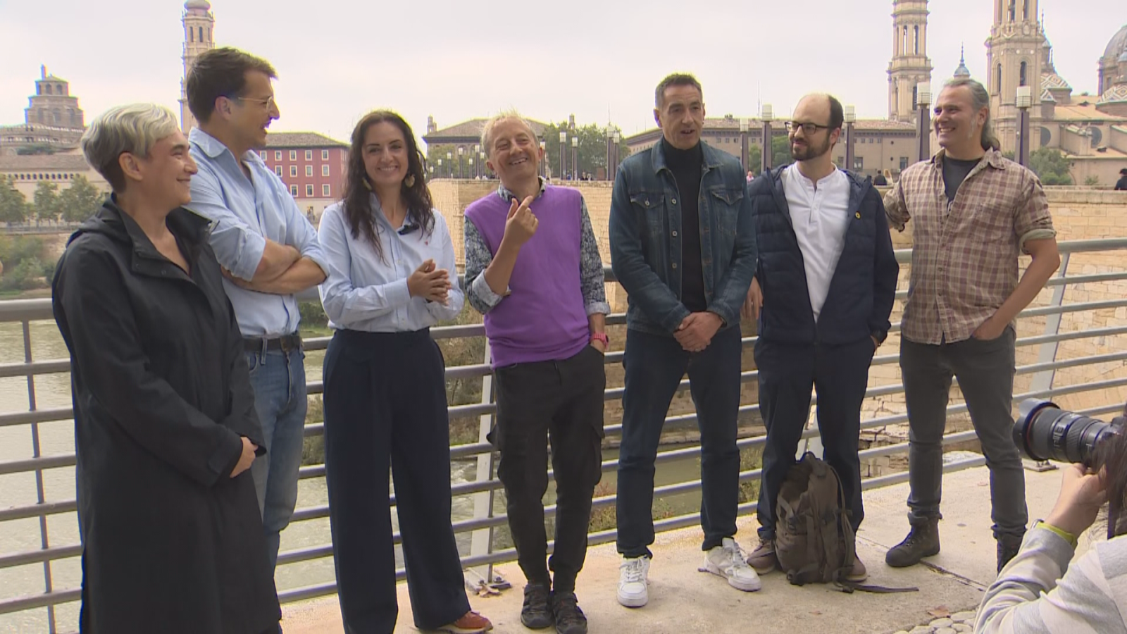 Siete actores de un programa cómico posan sonrientes junto al río Ebro en Zaragoza con la Basílica del Pilar al fondo y un fotógrafo en primer plano.