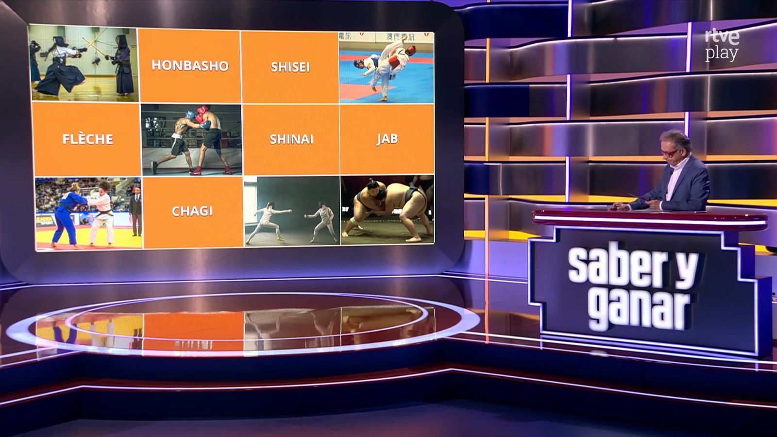 Plató de televisión con el presentador Jordi Hurtado y una gran pantalla que muestra un panel de juego sobre artes marciales y deportes de combate.