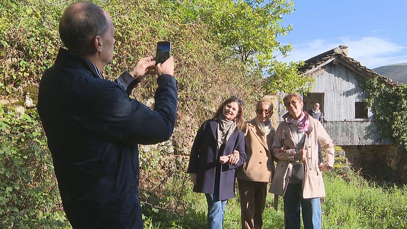 Un hombre fotografía a tres mujeres sonrientes que posan juntas sobre la hierba frente a una tradicional cabaña pasiega de piedra y madera en un día soleado.