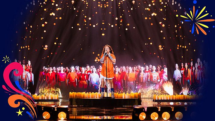 Joven cantante con camiseta naranja actúa en un escenario iluminado por velas y una pantalla gigante con un coro de niños.