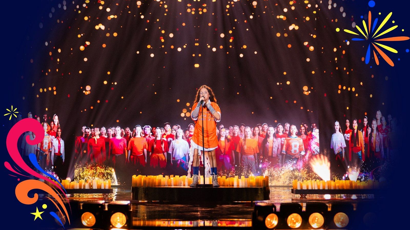 Joven cantante con camiseta naranja actúa en un escenario iluminado por velas y una pantalla gigante con un coro de niños.