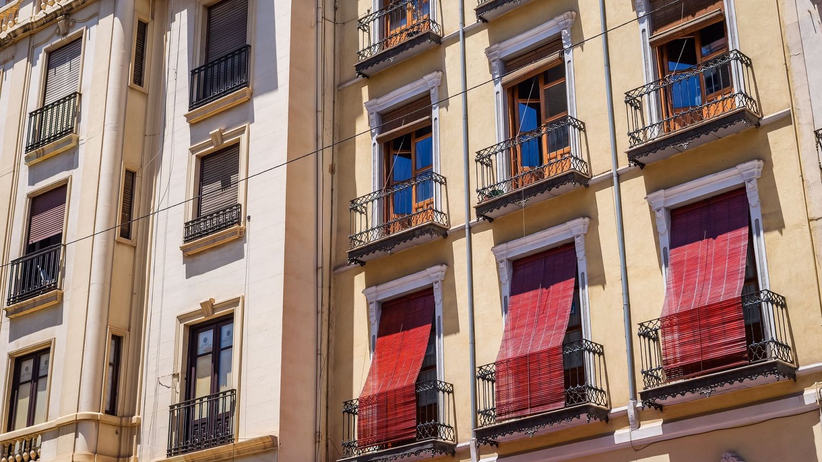 Fachadas soleadas de edificios residenciales tradicionales con balcones de hierro y persianas de madera en una jornada calurosa.