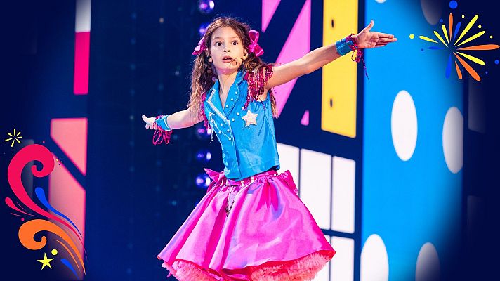Una niña cantante con chaleco azul brillante y falda fucsia actúa en un escenario iluminado con pantallas geométricas coloridas.