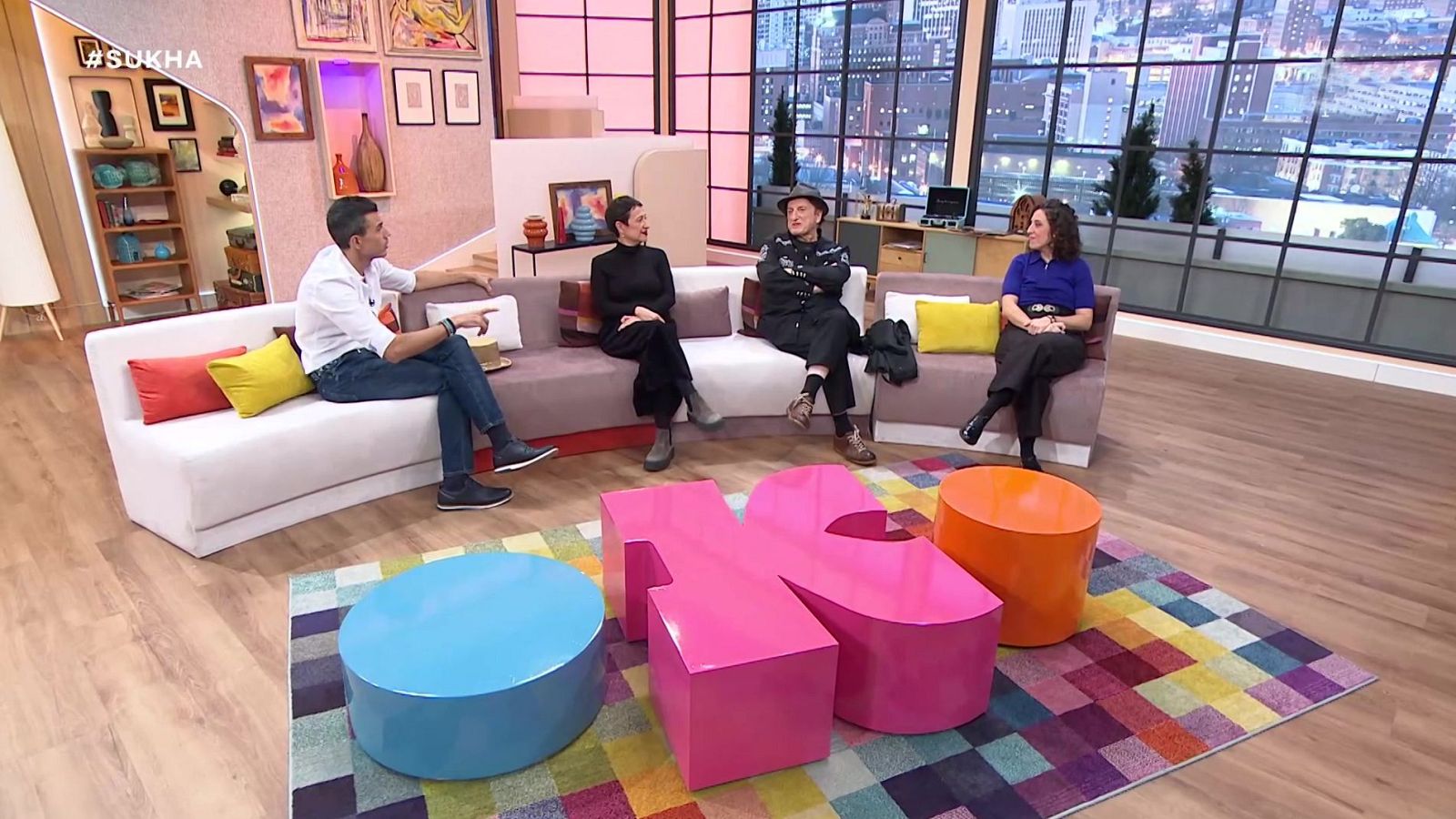 Plató de televisión luminoso y contemporáneo con cuatro personas conversando en un sofá modular curvado frente a mesas bajas de colores.