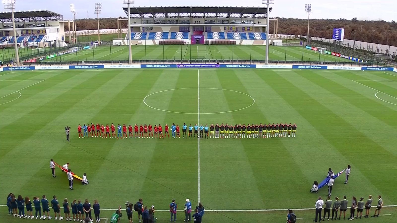 Vista panorámica de un campo de fútbol durante una ceremonia previa con jugadoras alineadas, árbitros y cuerpos técnicos en el césped.