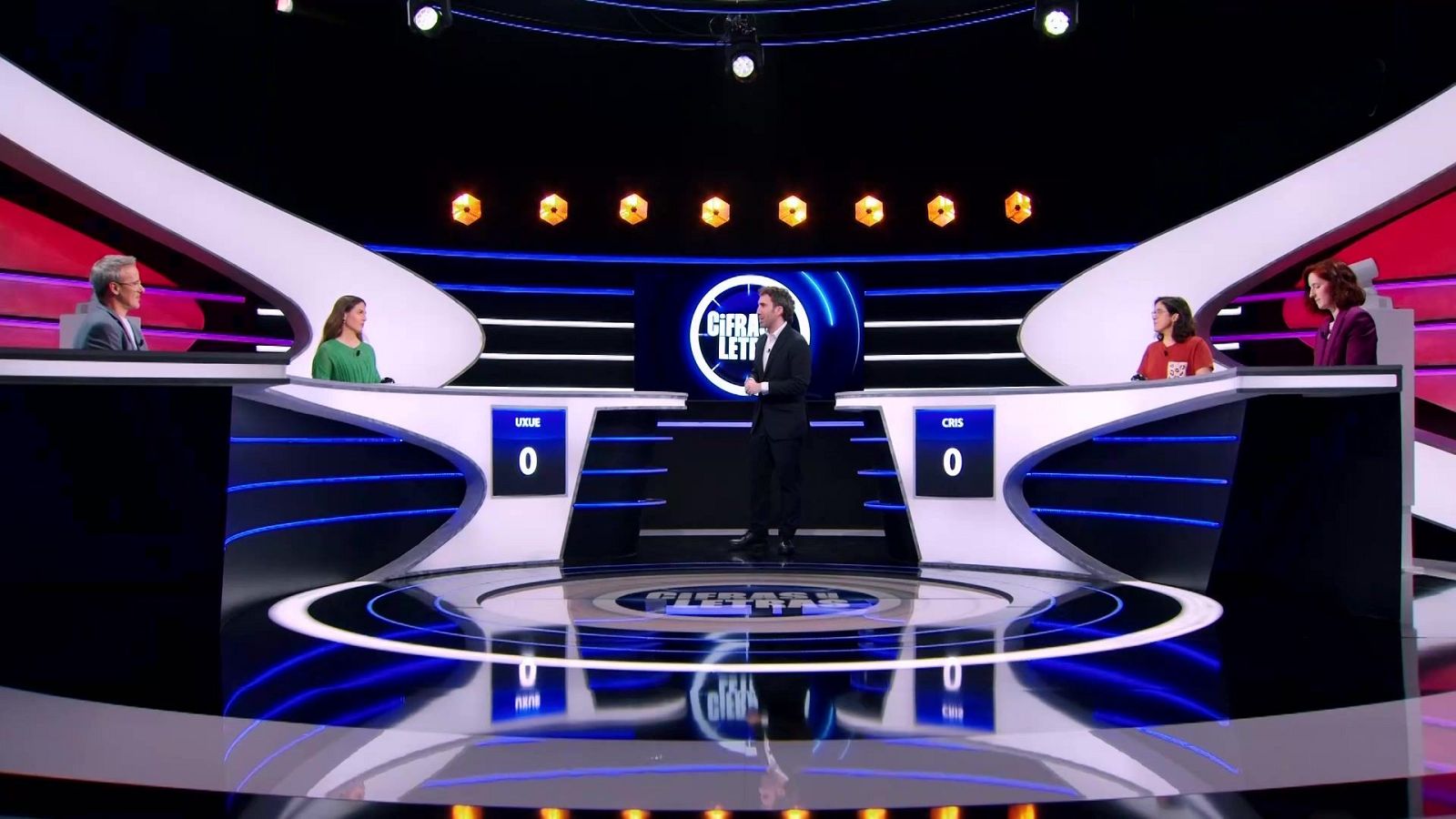Plató de televisión del concurso Cifras y letras con el presentador en el centro, los concursantes Uxue y Cris en los atriles y los colaboradores a los lados.