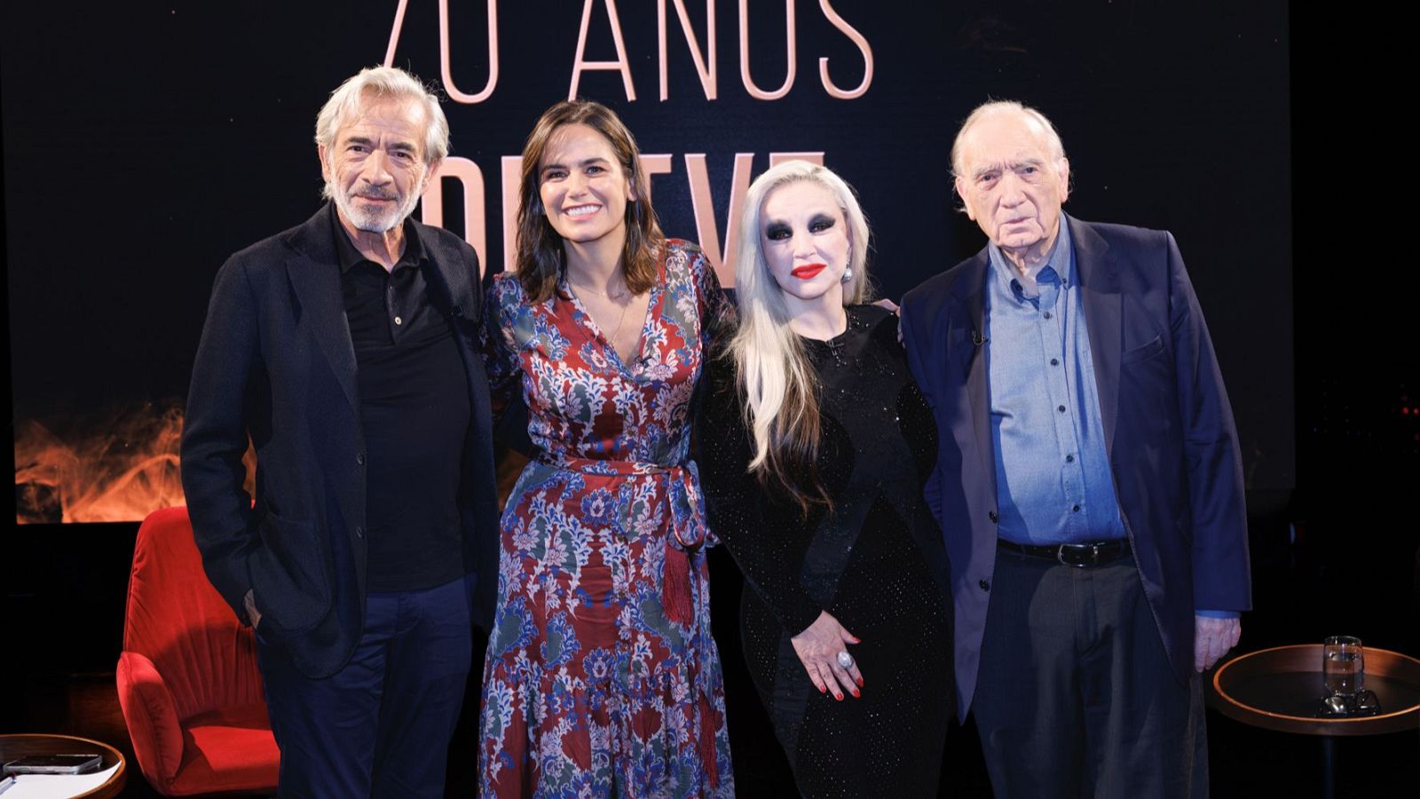 Cuatro figuras de la cultura española posan sonrientes en un plató con fondo oscuro e iluminación suave frente a una pantalla conmemorativa luminosa.