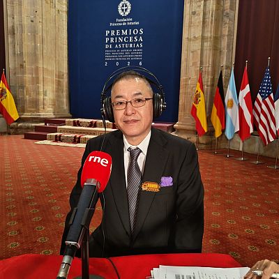 Hombre de mediana edad con gafas y traje formal sentado ante un micrófono de RNE durante una entrevista en un salón institucional con banderas.