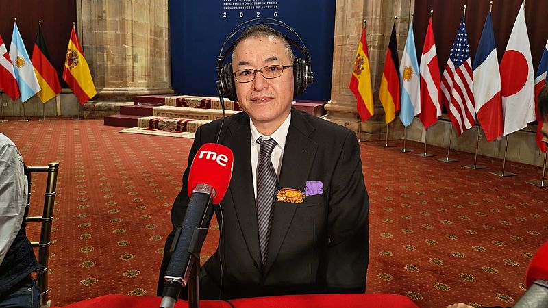 Hombre de mediana edad con gafas y traje formal sentado ante un micrófono de RNE durante una entrevista en un salón institucional con banderas.