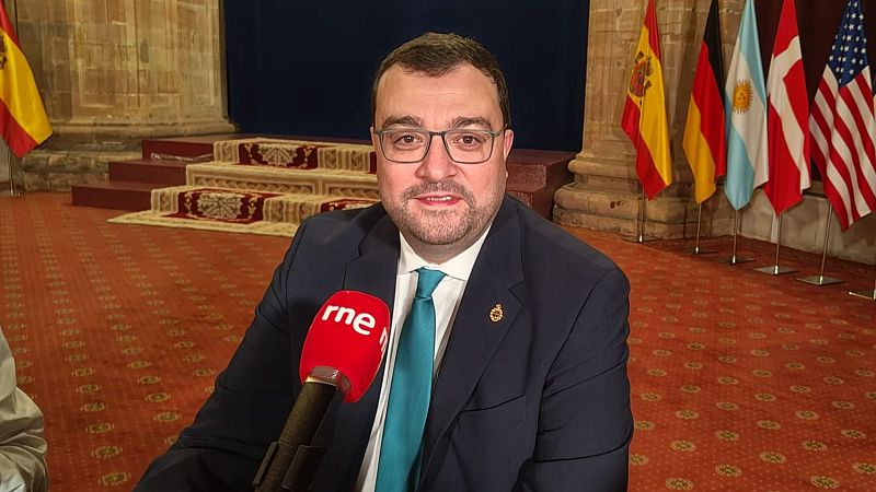 Adrián Barbón ofrece una entrevista en un salón noble decorado con banderas y muros de piedra, luciendo americana azul y una insignia en la solapa.