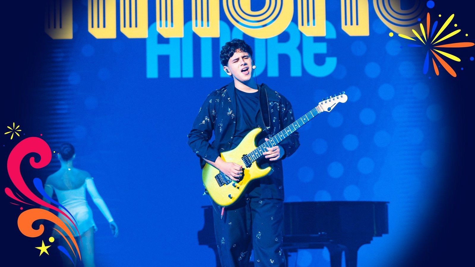 Joven cantante con guitarra eléctrica amarilla actúa en un escenario iluminado de azul con la palabra AMORE proyectada al fondo.