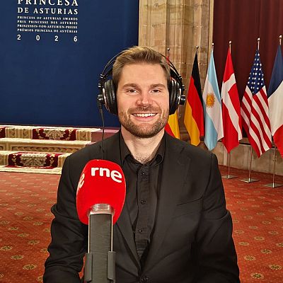 Un locutor sonriente con auriculares y micrófono de RNE en los Premios Princesa de Asturias, con un panel oficial y banderas de fondo.