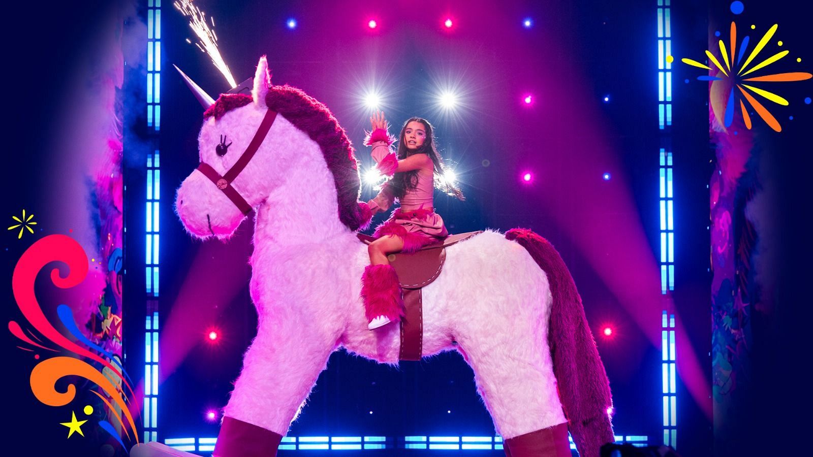 Una niña con atuendo fucsia actúa sobre un enorme unicornio de peluche blanco con chispas pirotécnicas en el cuerno, en un escenario iluminado con luces de neón.