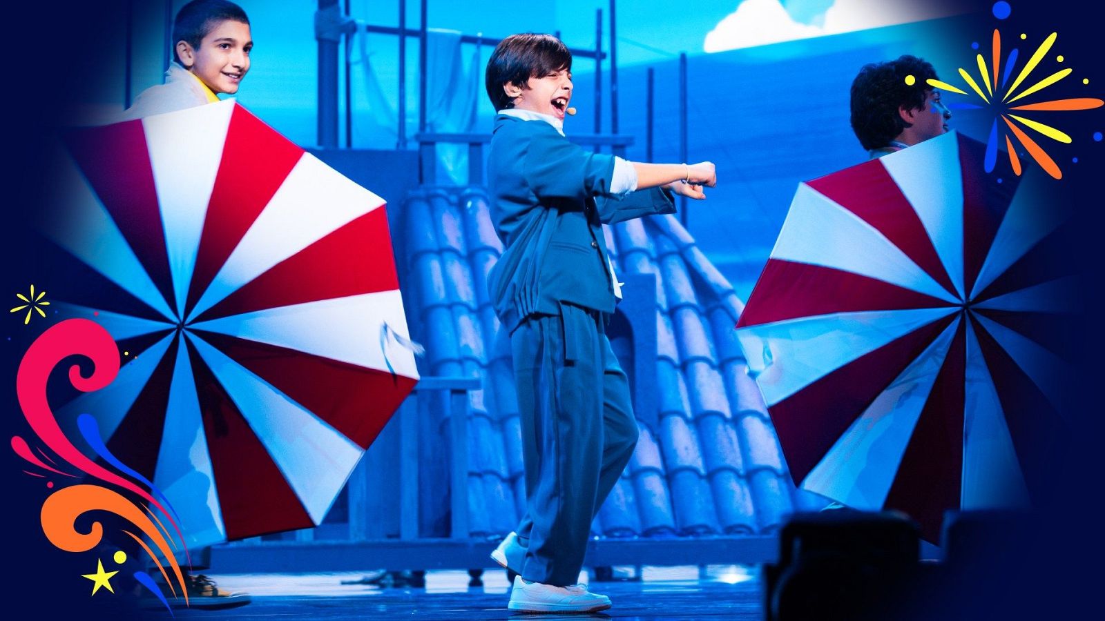 Actuación musical de un joven cantante con traje azul y zapatillas blancas junto a bailarines con paraguas rojo y blanco en un escenario con escenografía de tejado y luces azules.