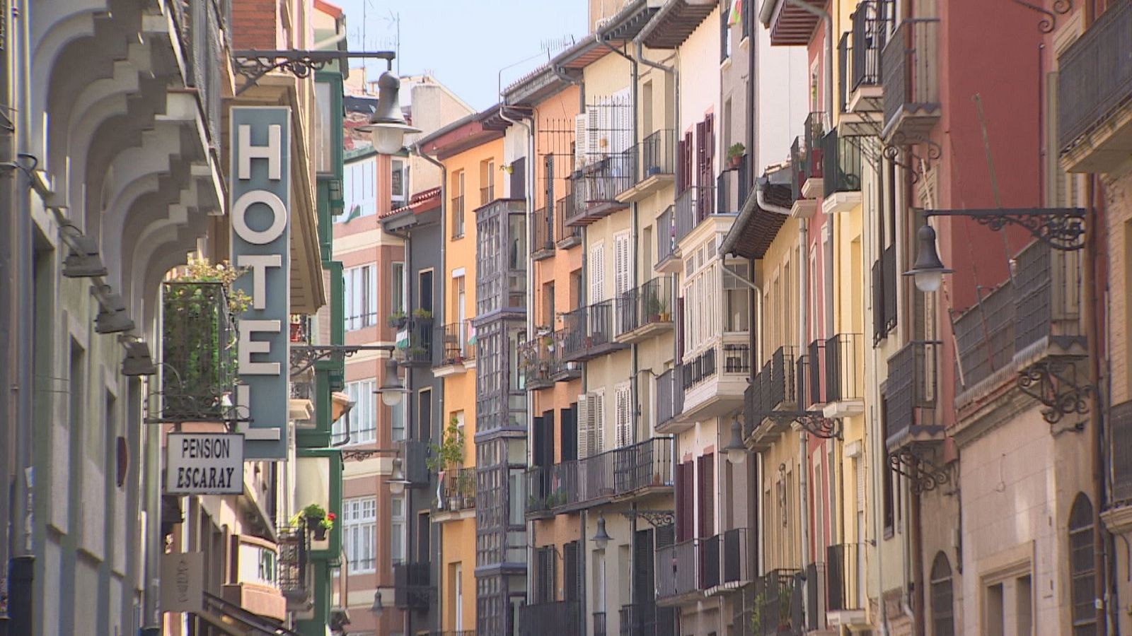Vista de una calle estrecha y tradicional con edificios de tonos cálidos, balcones de forja y carteles de un hotel y pensión en primer plano.