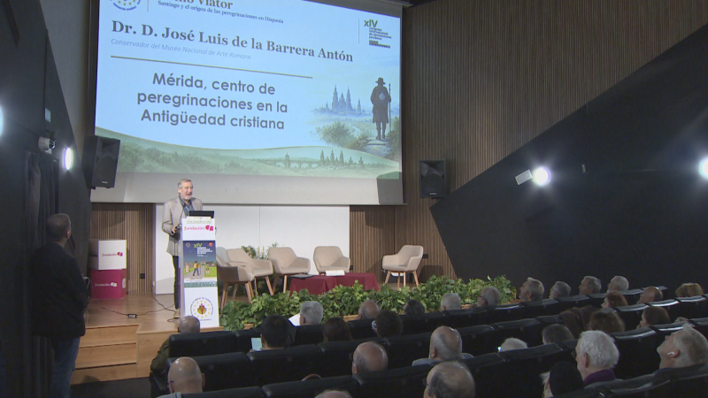 En un auditorio lleno, un ponente interviene en el estrado frente a una gran pantalla que proyecta el título 'Mérida, centro de peregrinaciones en la Antigüedad cristiana'.