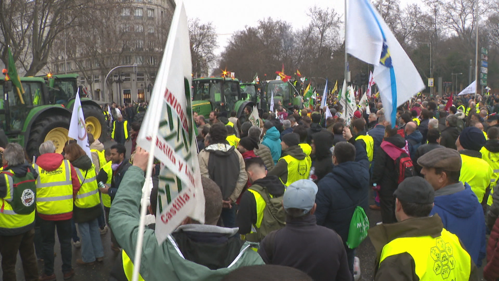 Manifestación de agricultores con tractores y banderas regionales que avanza por una amplia avenida urbana en invierno.