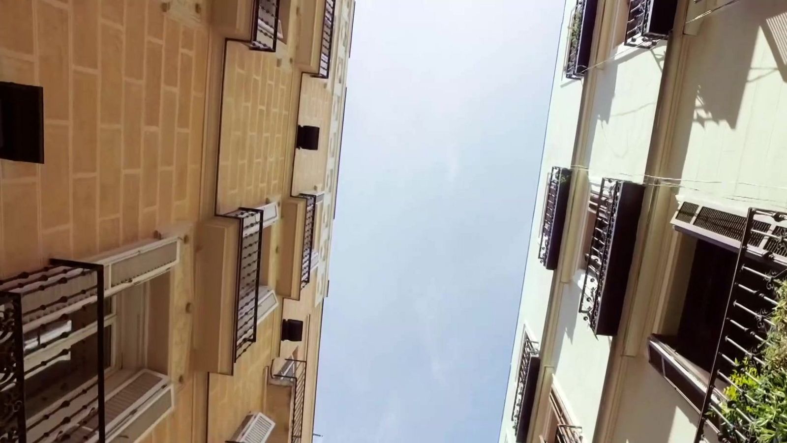 Vista en contrapicado de dos edificios con balcones de hierro forjado en una calle estrecha que enmarca el cielo.