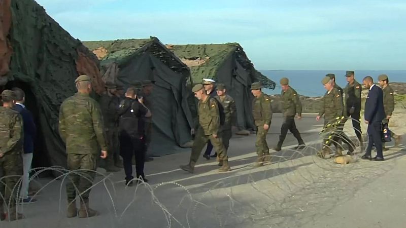 El Rey Felipe VI camina con uniforme de camuflaje junto a mandos militares en un campamento costero con tiendas y redes de camuflaje.
