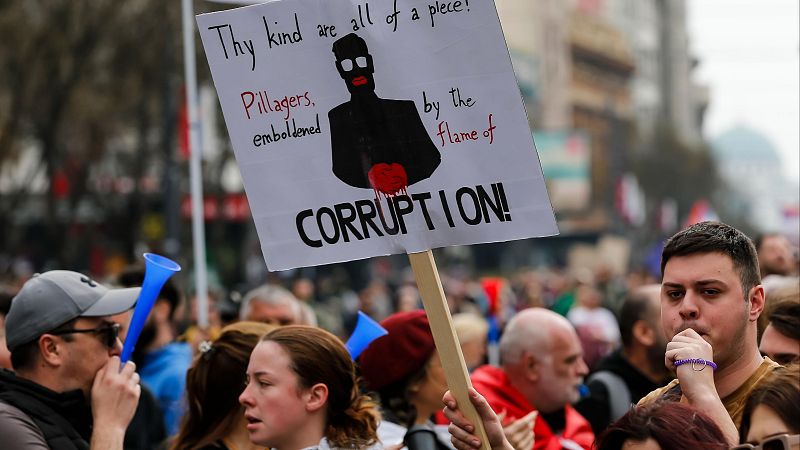 Manifestación en una avenida de Belgrado donde destacan una pancarta con la palabra corrupción y asistentes haciendo sonar silbatos.