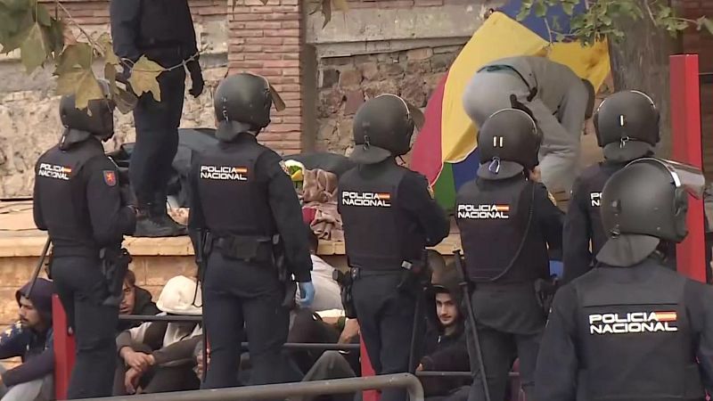 Agentes de la Policía Nacional forman una línea de seguridad frente a varias personas jóvenes sentadas en el suelo en un área urbana.