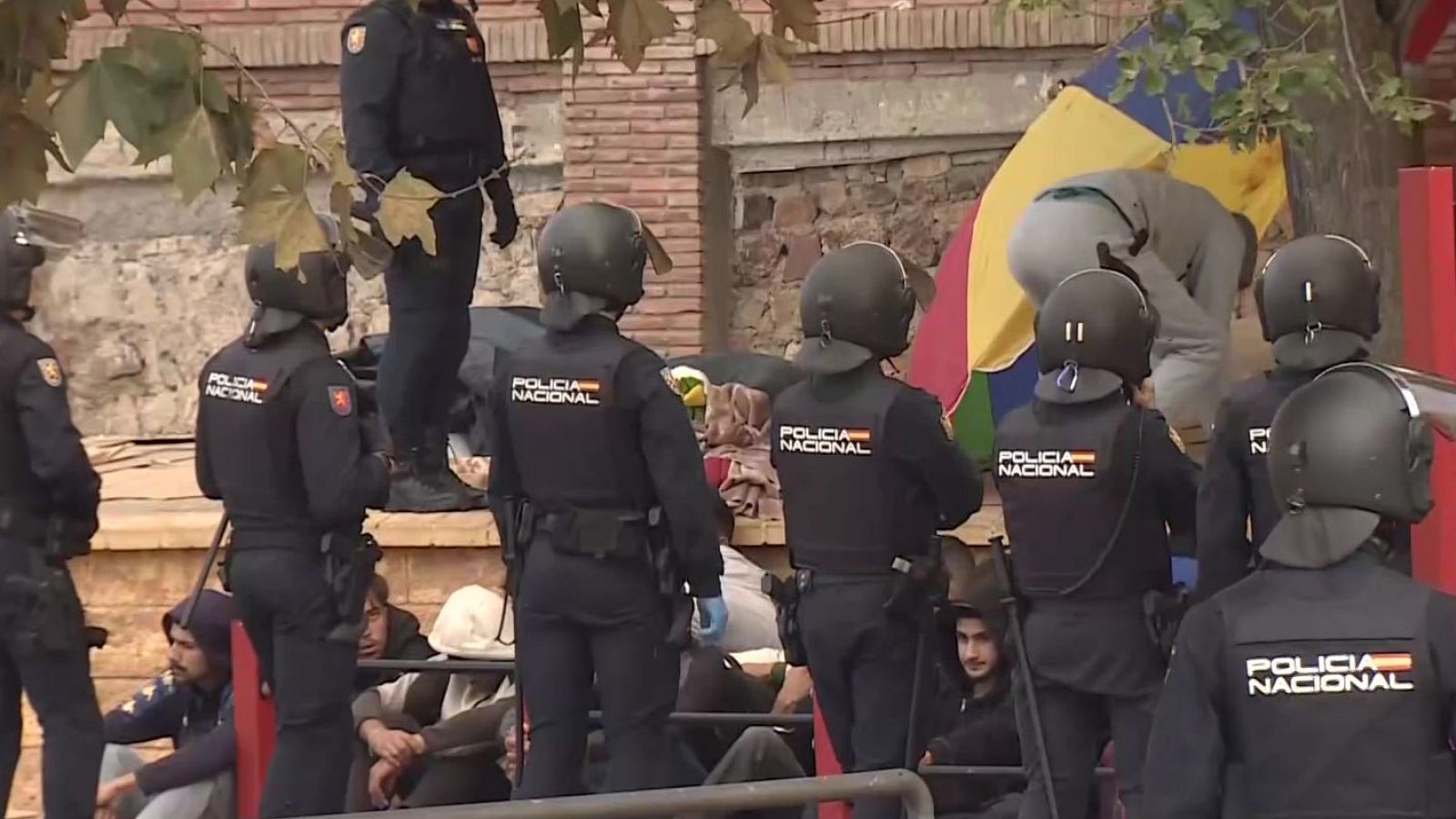Agentes de la Policía Nacional con equipamiento táctico vigilan a jóvenes sentados en el suelo junto a una barandilla en una zona urbana, con un campamento improvisado al fondo.