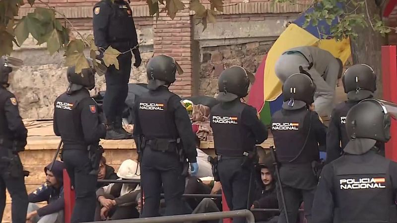 Agentes de la Policía Nacional con equipamiento táctico vigilan a jóvenes sentados en el suelo junto a una barandilla en una zona urbana, con un campamento improvisado al fondo.