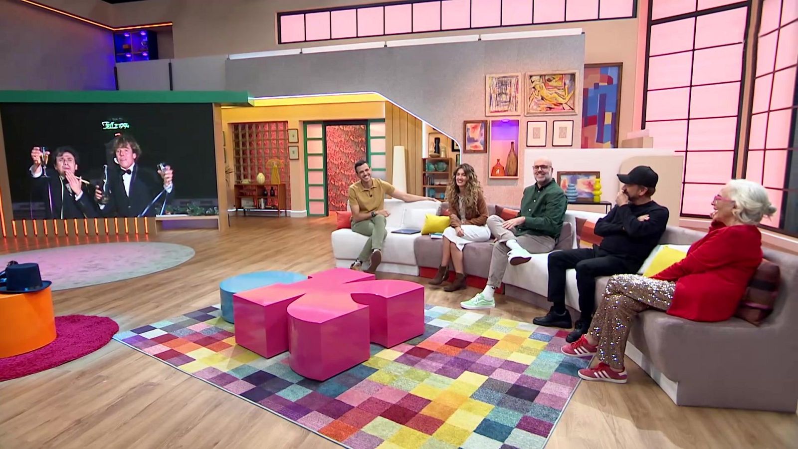 Cinco personas conversan sentadas en un sofá modular en un plató moderno con una mesa fucsia y una gran pantalla lateral.