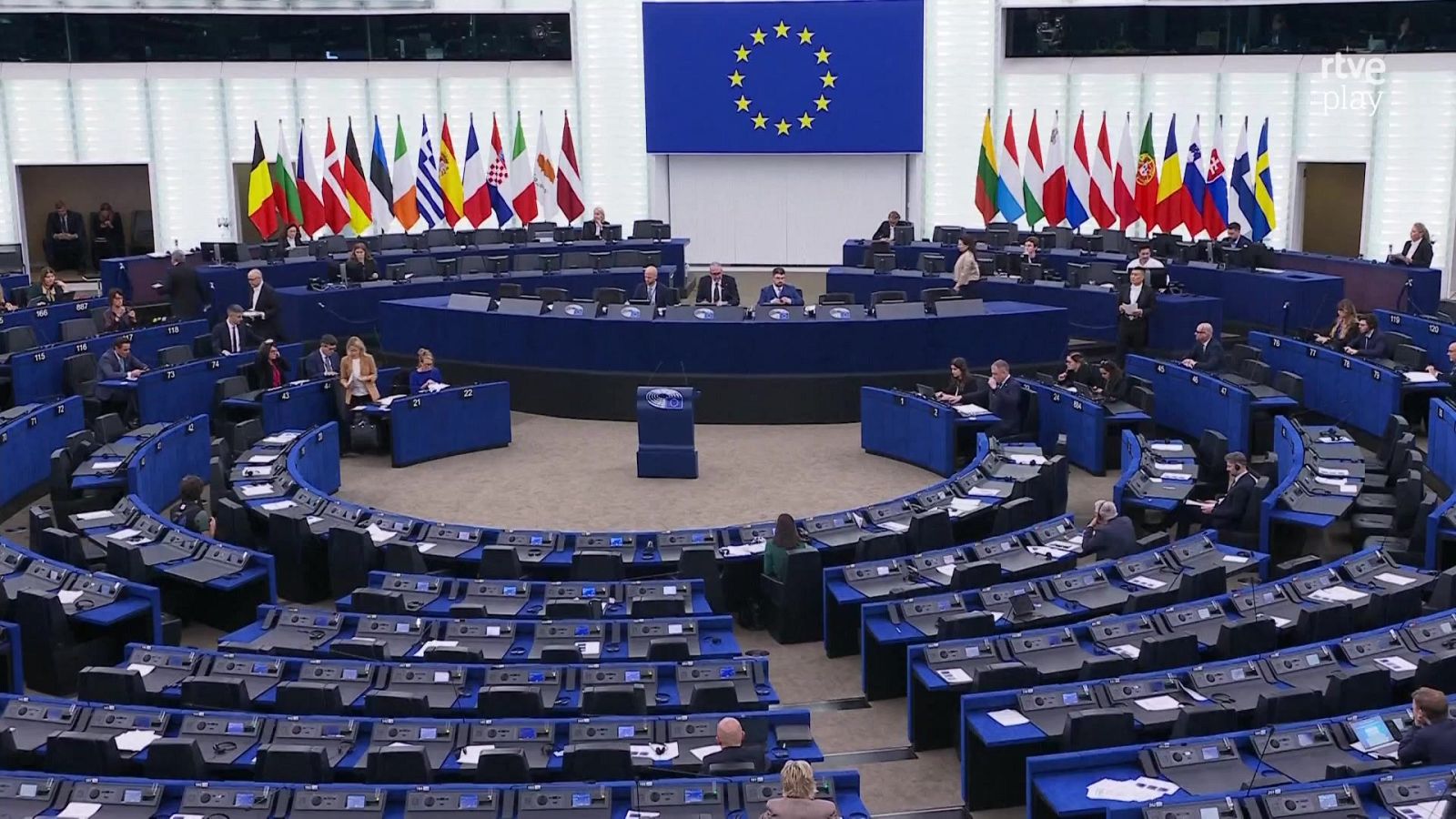 Vista panorámica del hemiciclo del Parlamento Europeo con escaños semicirculares, bandera de la UE al fondo y el logo de rtve play en la esquina superior derecha.