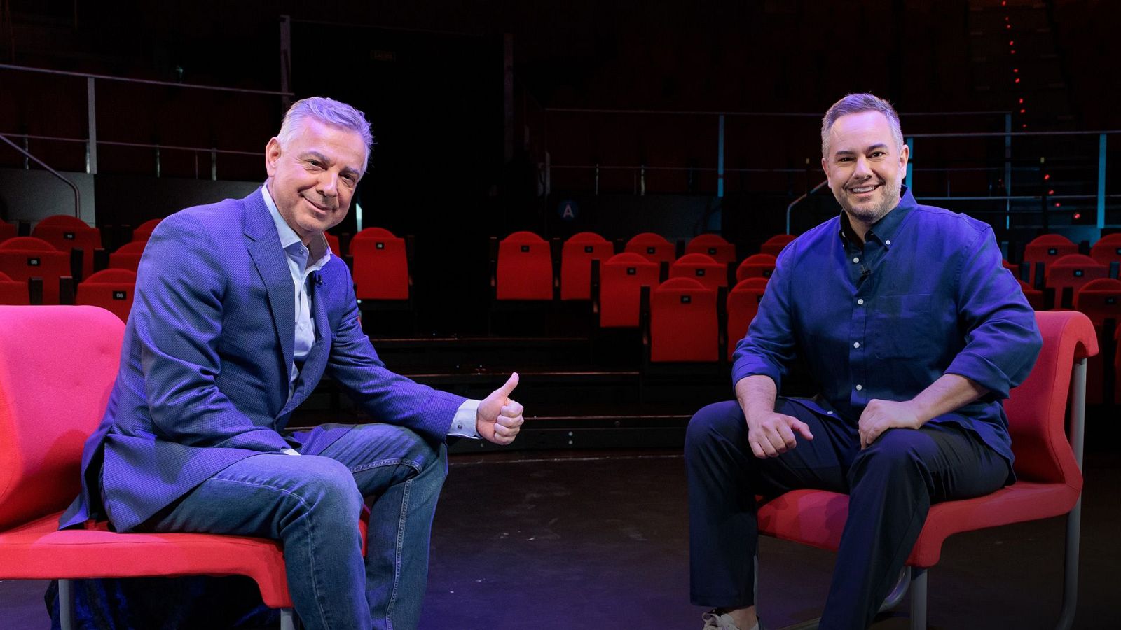 Dos hombres sentados en butacas rojas sobre el escenario de un teatro vacío, sonriendo y mirando a la cámara.
