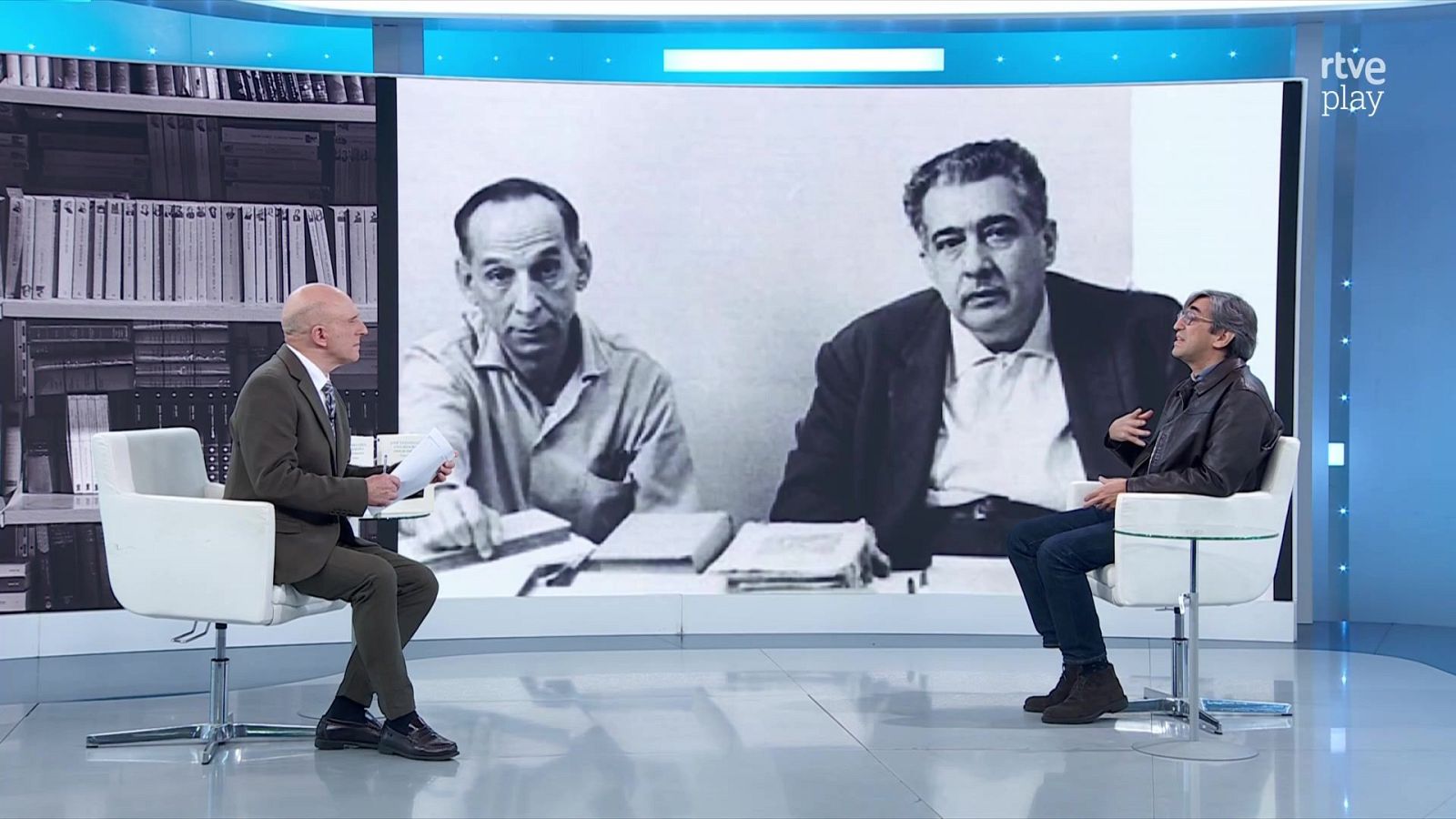 Dos personas conversan sentadas en butacas blancas en un plató de televisión, con una gran pantalla curva al fondo que proyecta imágenes de archivo.