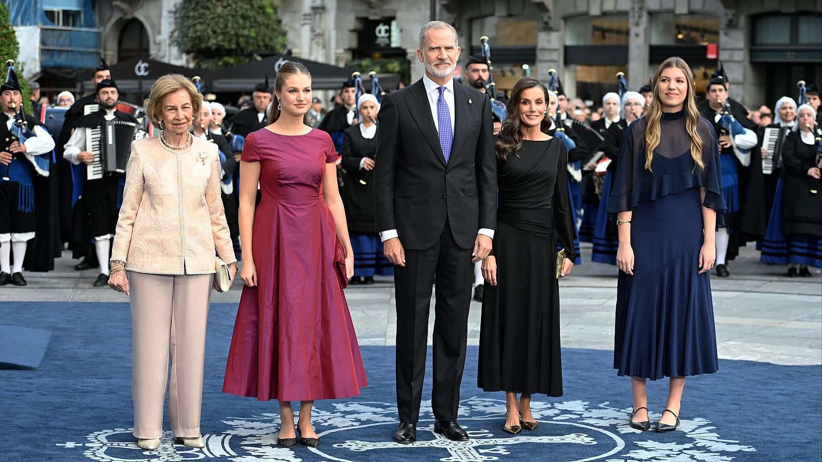 La Familia Real española posa al completo sobre una alfombra azul en Oviedo, acompañada de músicos tradicionales con gaitas y rodeada de público.