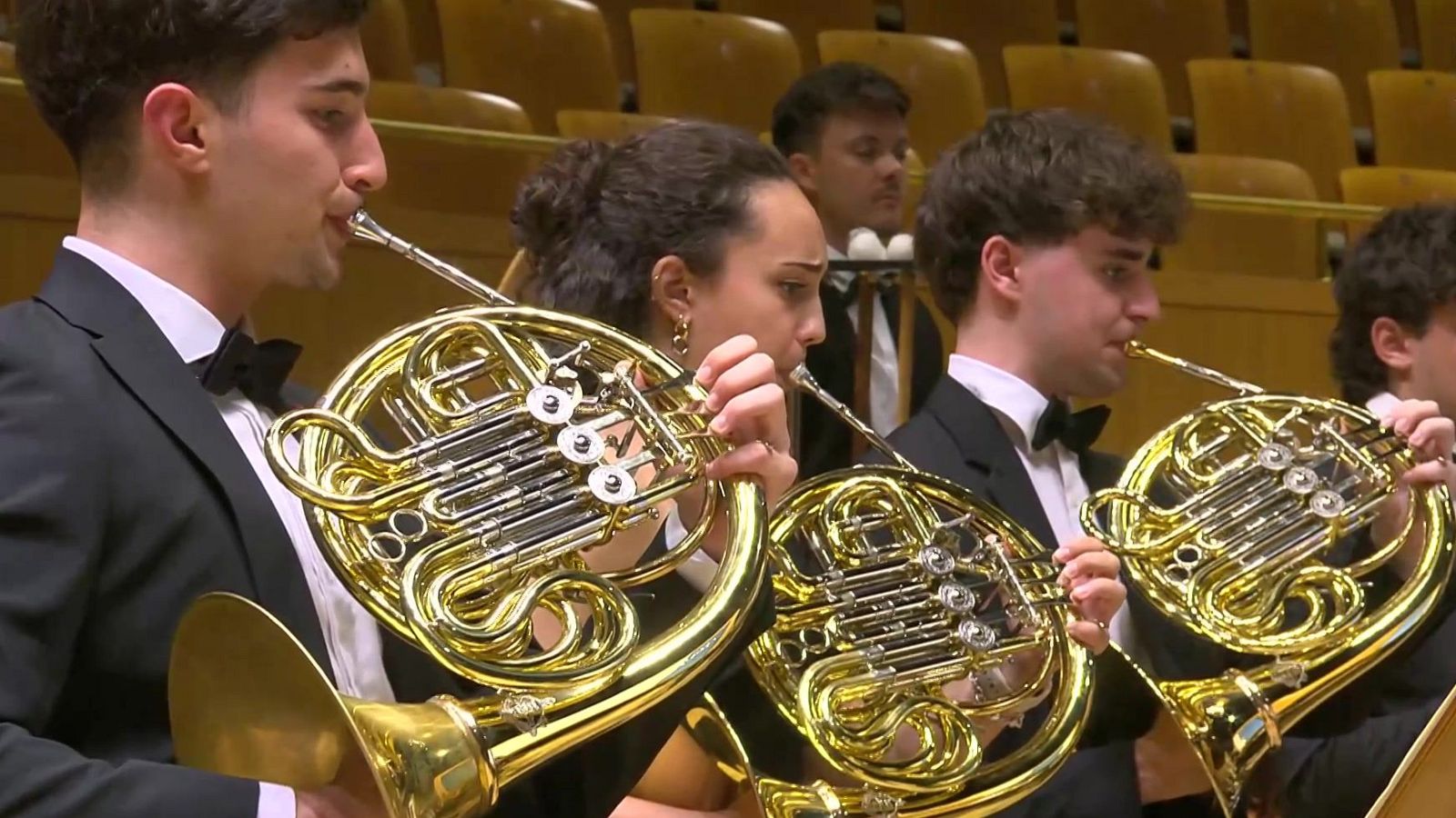 Jóvenes músicos de una orquesta sinfónica interpretan una obra clásica tocando la trompa en el escenario de un auditorio bajo la emisión de Los conciertos de La 2.