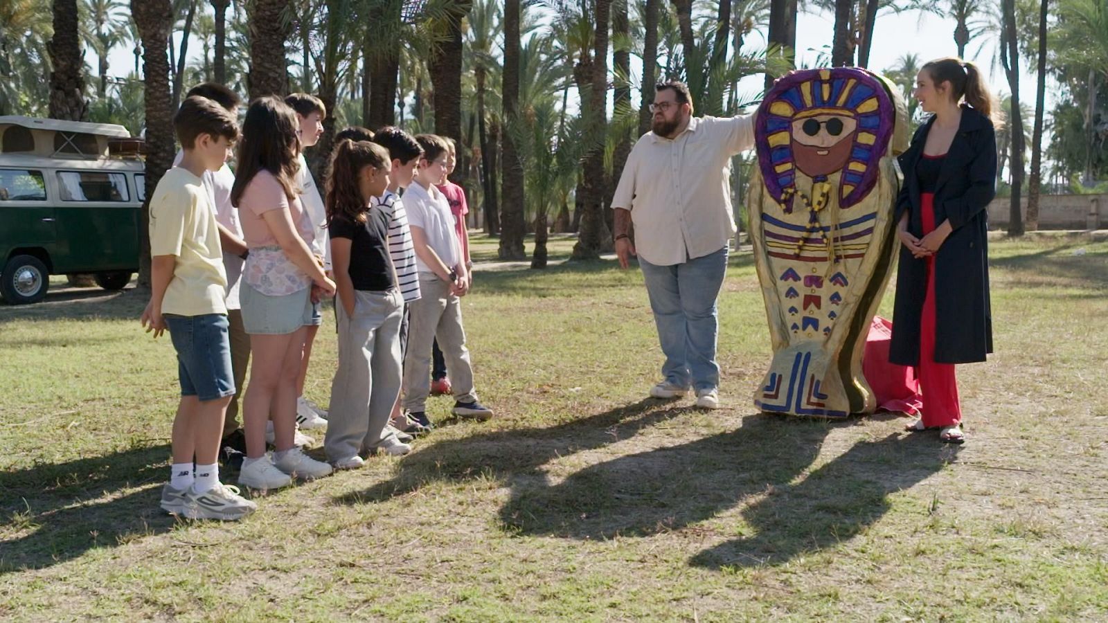 Grupo de escolares y dos educadores interactúan junto a una maqueta de sarcófago egipcio al aire libre, rodeados de palmeras y con una furgoneta al fondo.
