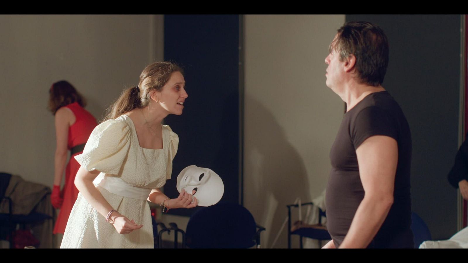 Una actriz con vestido blanco y máscara teatral ensaya una escena dramática frente a su compañero en una sala.