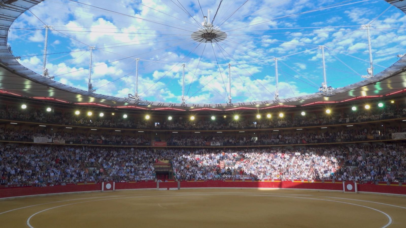 Vista panorámica de la Plaza de Toros con tendidos llenos de público, ruedo de albero y una cubierta de estructura tensada bajo un cielo azul.