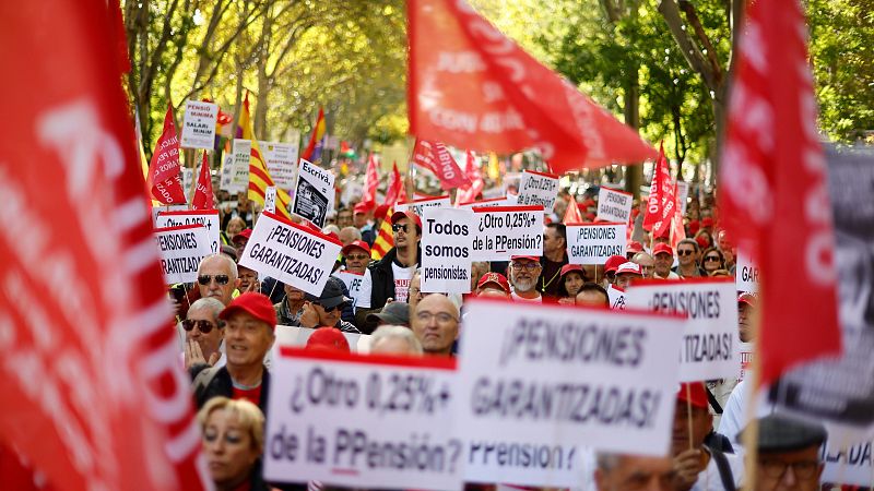 Manifestación masiva de pensionistas con pancartas y banderas rojas avanzando por una avenida arbolada.