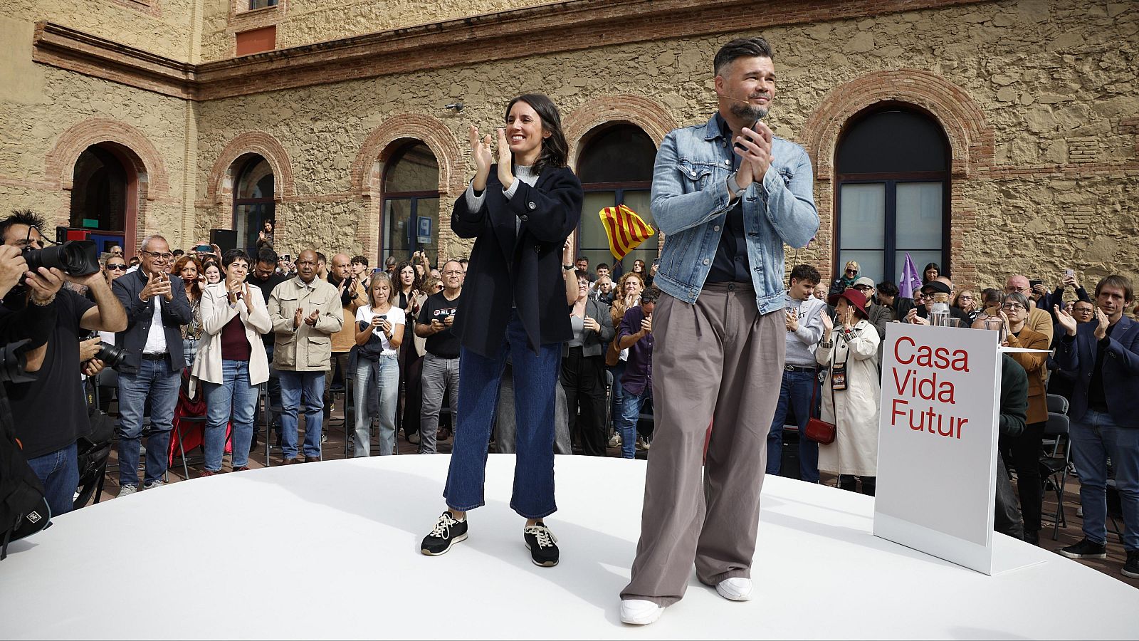 Irene Montero y Gabriel Rufián aplauden sonrientes sobre una tarima ante un público numeroso y fotógrafos, junto a un atril con la inscripción 'Casa / Vida / Futur'.