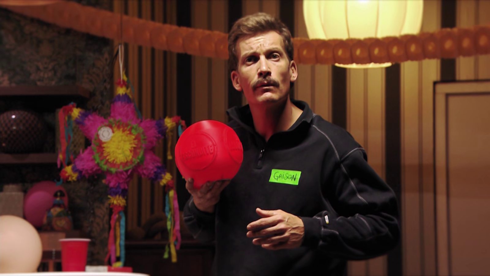 El presentador Grison sostiene una pelota roja y gesticula con la mano en un salón decorado con una piñata multicolor y guirnaldas.