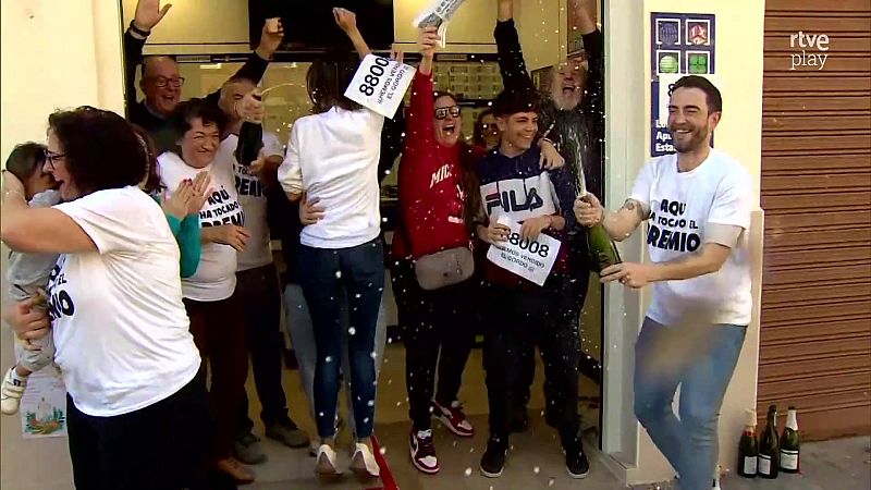 Un grupo celebra con champán y carteles del número premiado 88008 frente a una administración de lotería.