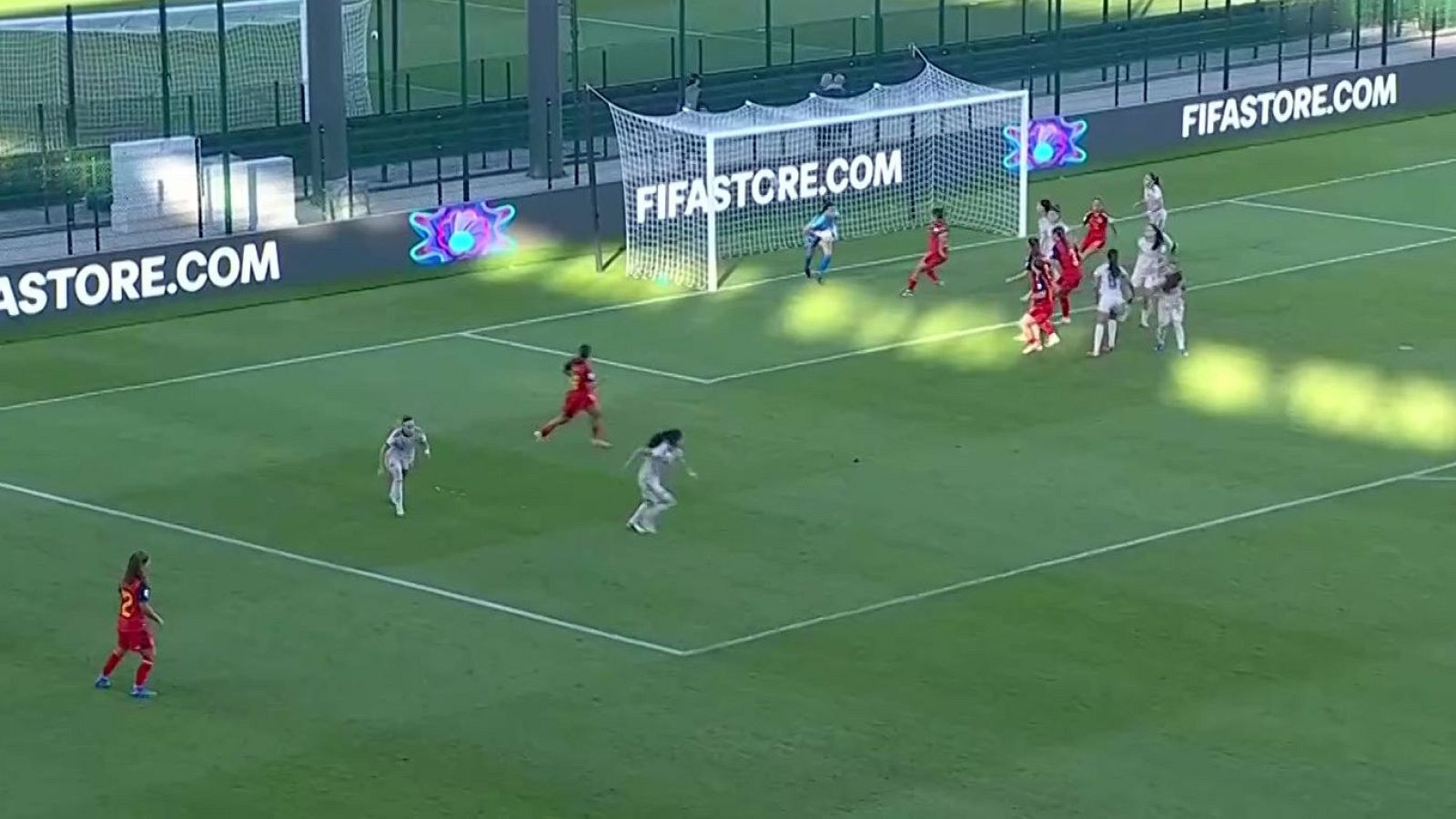 Vista picada de una jugada de ataque en un partido de fútbol femenino con futbolistas de equipos rojo y blanco en el área.