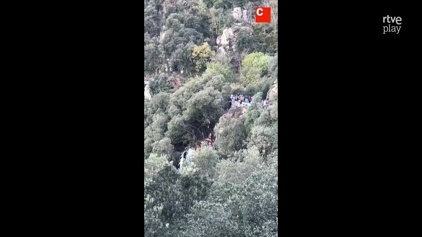 Emisión televisiva que muestra una ladera montañosa escarpada con vegetación y personas observando una zona de posible rescate.