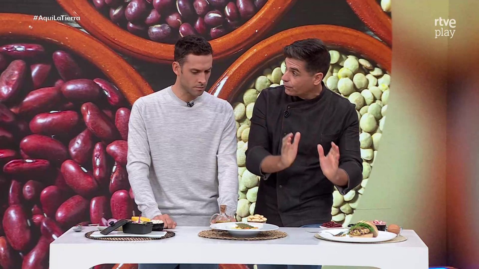 Dos hombres cocinan en un plató de televisión ante una mesa con platos de legumbres, acompañados por una gran pantalla al fondo.