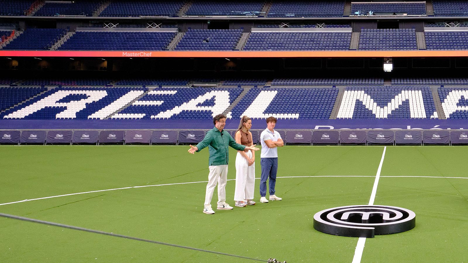 Los tres jueces de MasterChef, Pepe Rodríguez, Samantha Vallejo-Nágera y Jordi Cruz, están de pie sobre el césped del Estadio Santiago Bernabéu junto al logotipo del programa.