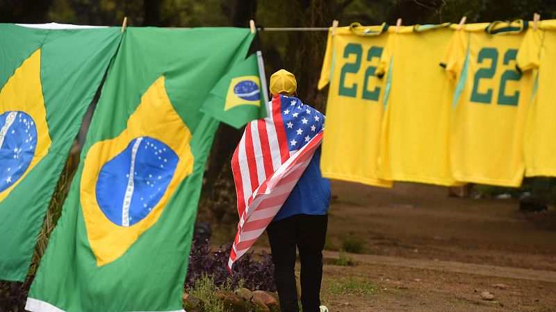 Persona de espaldas con la bandera de Estados Unidos camina por un sendero junto a un tendedero con banderas de Brasil y camisetas amarillas con el número 22.