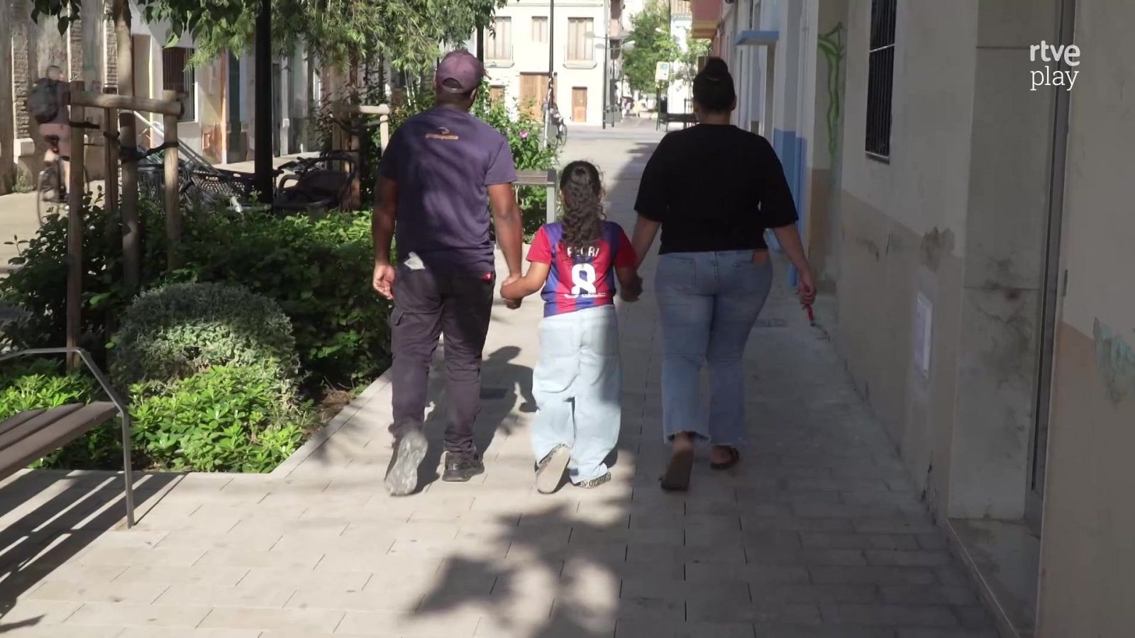 Tres personas caminan de espaldas cogidas de la mano por una calle peatonal, destacando una niña en el centro con camiseta del Barça y el nombre de Pedri en la espalda.