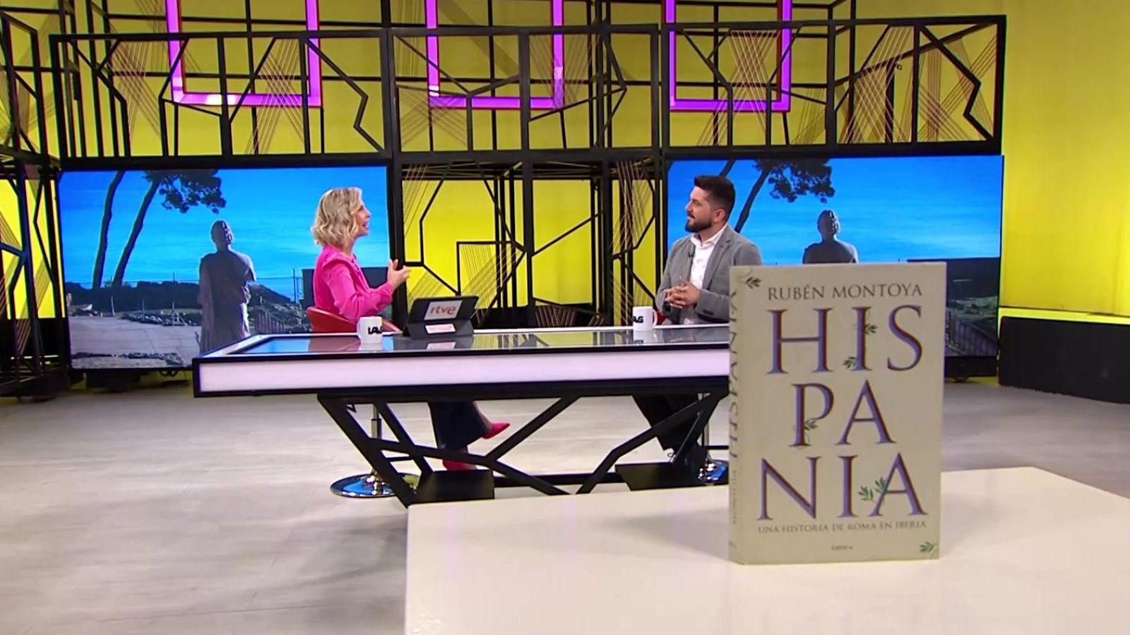 La presentadora Mara Peterssen entrevista al historiador Rubén Montoya en el plató del programa La aventura del Saber, con un libro en primer plano.